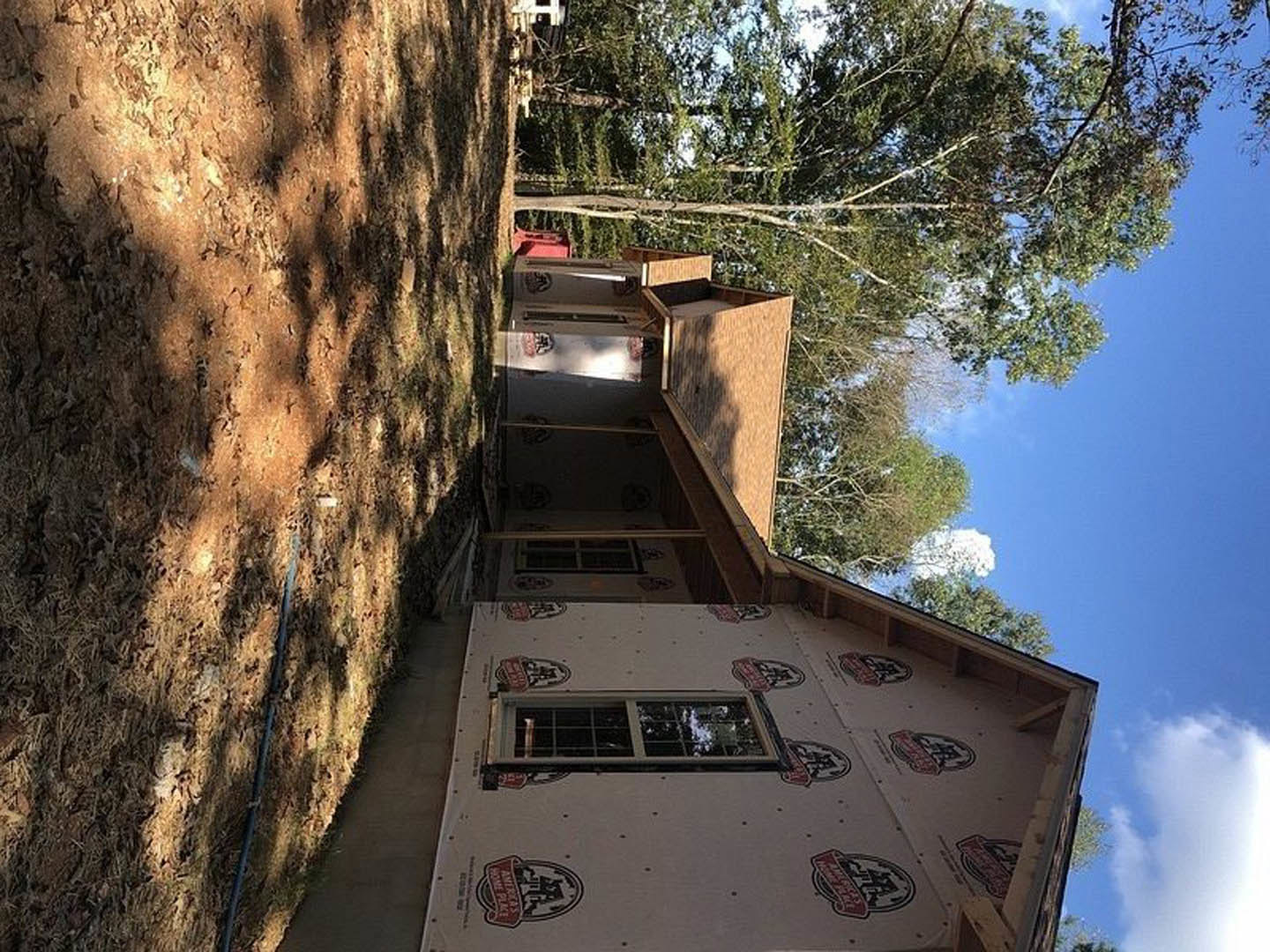 Partially built house with white foam insulation on exterior walls, white-framed window, dirt yard, blue hose, black pipe, and trees in the background.