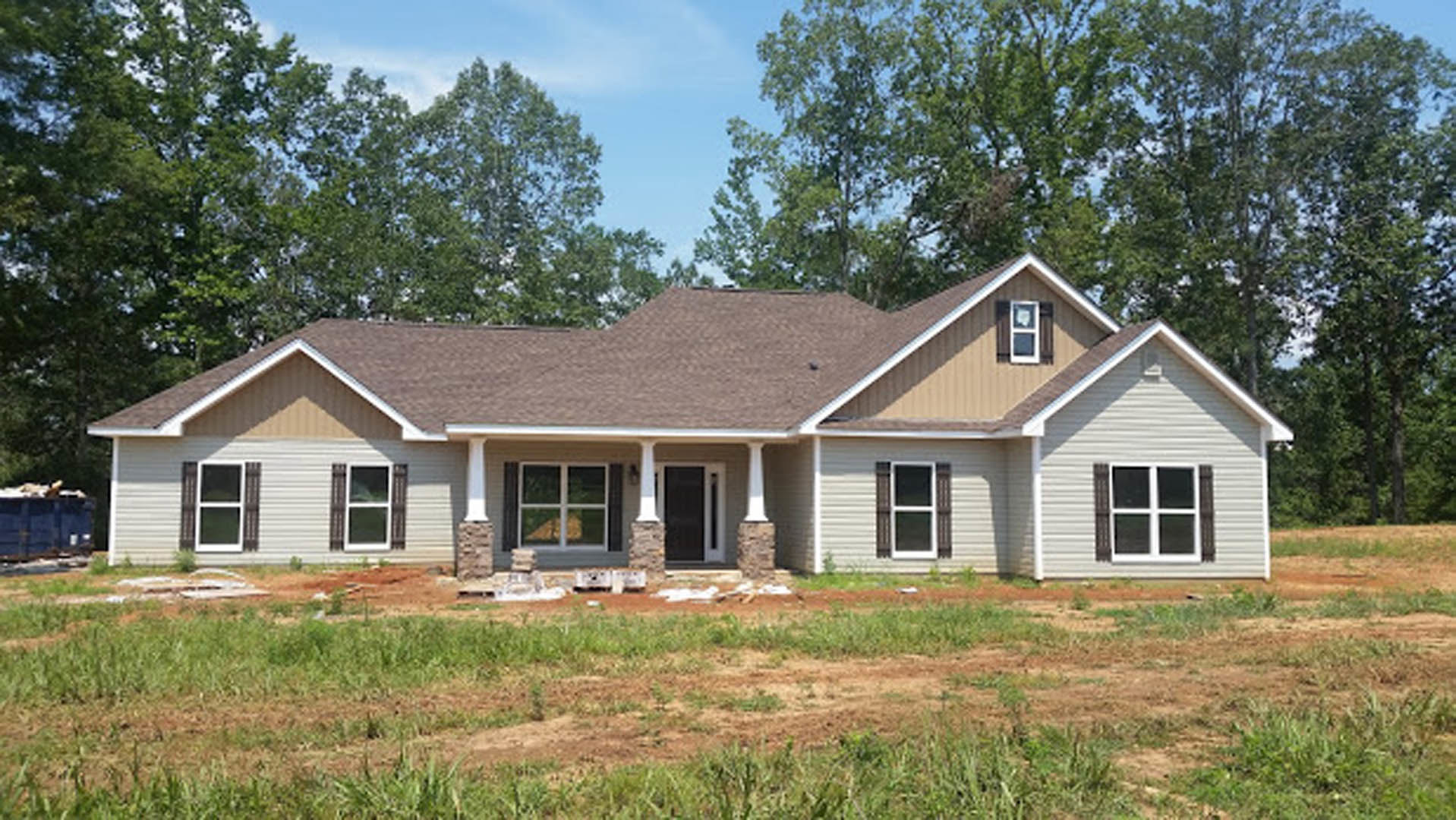 Partially built house with brown roof, white-framed window, exposed wood framing, surrounded by grass and dirt, mature trees in background