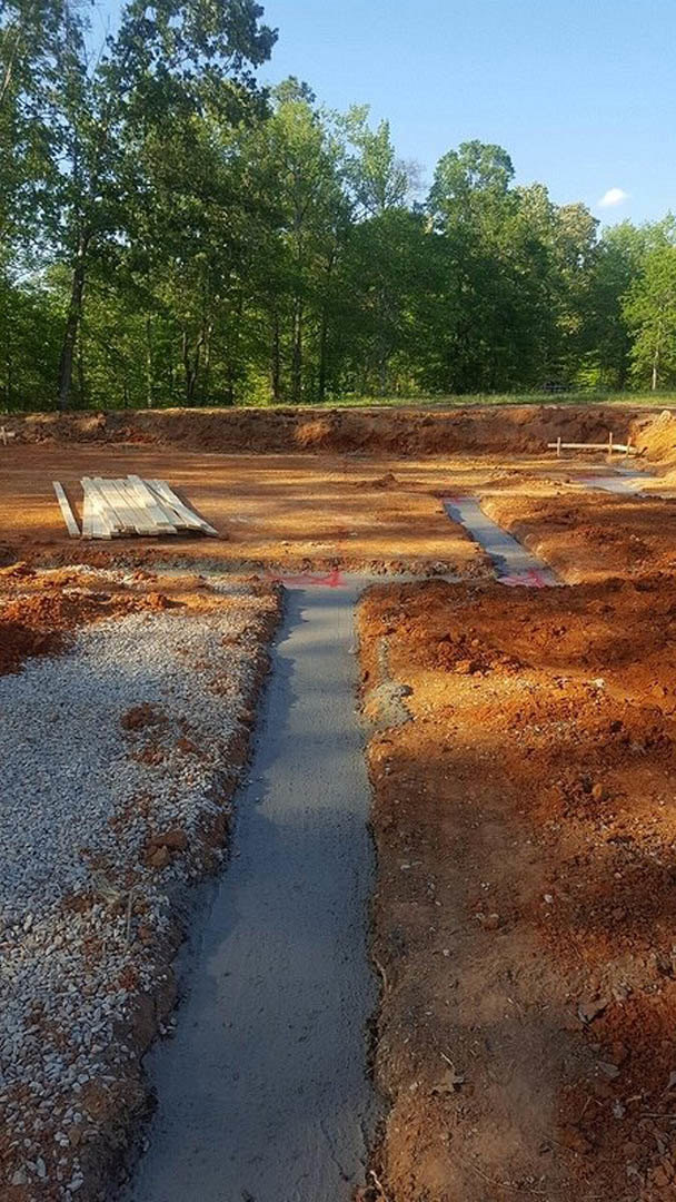 Construction site with exposed dirt trench, stacked wood planks, gravel surface, and surrounding trees under open sky