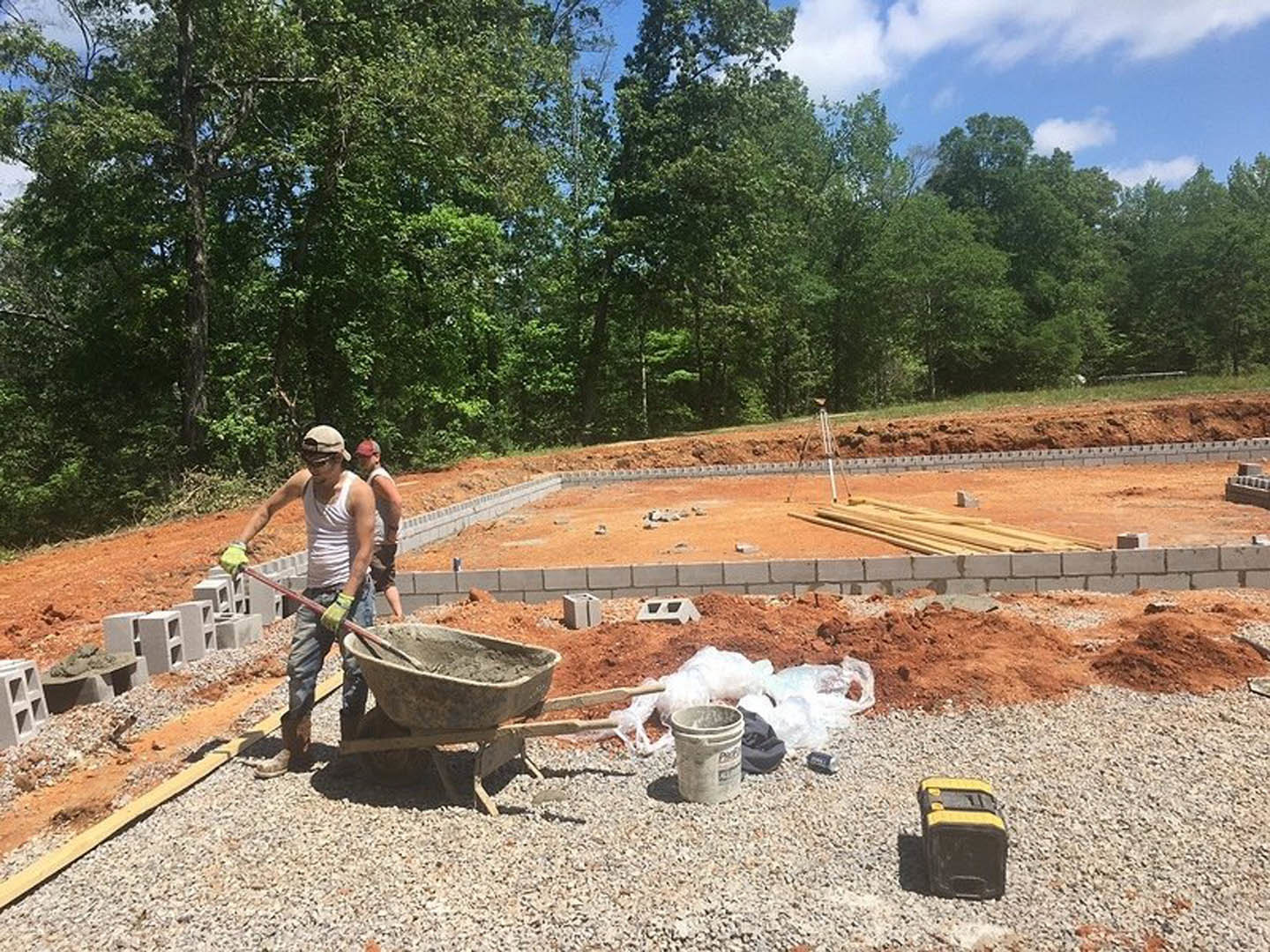 Two men building a home foundation, one holding a red rod near a yellow and black tool, cement bucket and bowl on soil, trees and blue sky in background