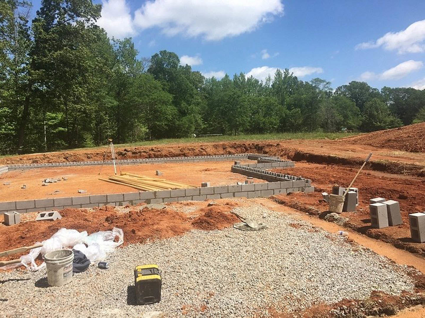 Construction site with exposed soil, scattered tools, and a few mature trees under a partly cloudy blue sky