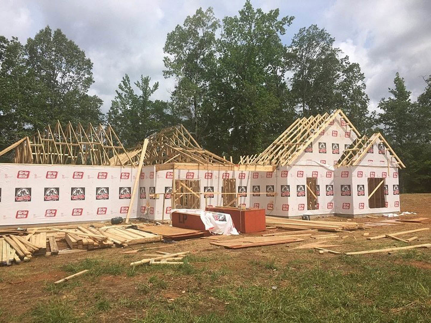 Wood-framed house under construction with exposed beams, surrounded by grass and trees under a cloudy sky