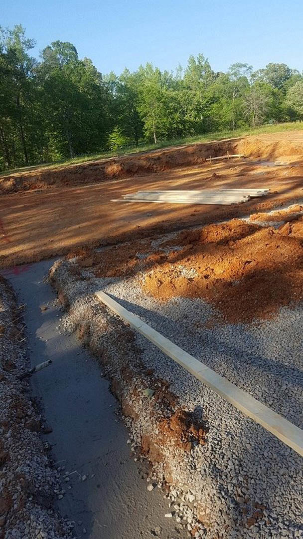 Wood planks and gravel at a residential construction site, dirt trench in foreground, trees and blue sky in background
