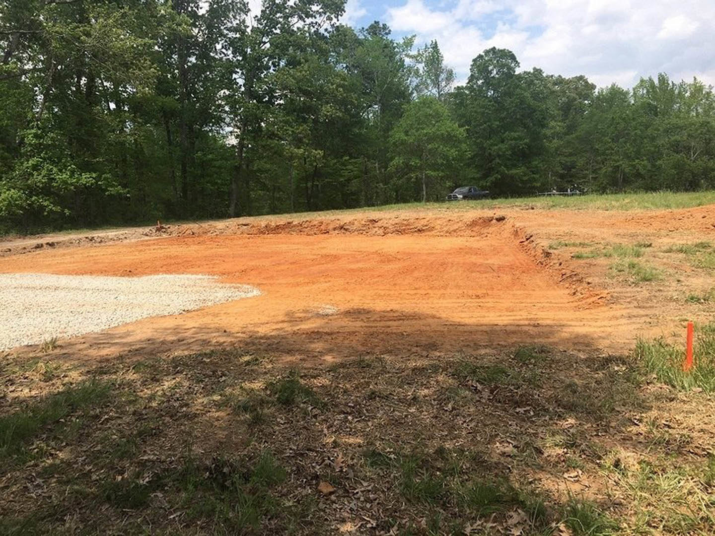 Dirt field with scattered grass and soil, bordered by leafy trees under a partly cloudy sky