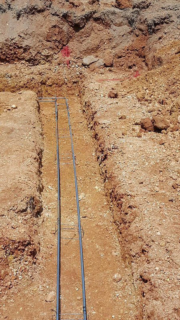 Freshly dug trench in brown soil outdoors, exposed pipe partially visible along the bottom, loose dirt piled on either side.
