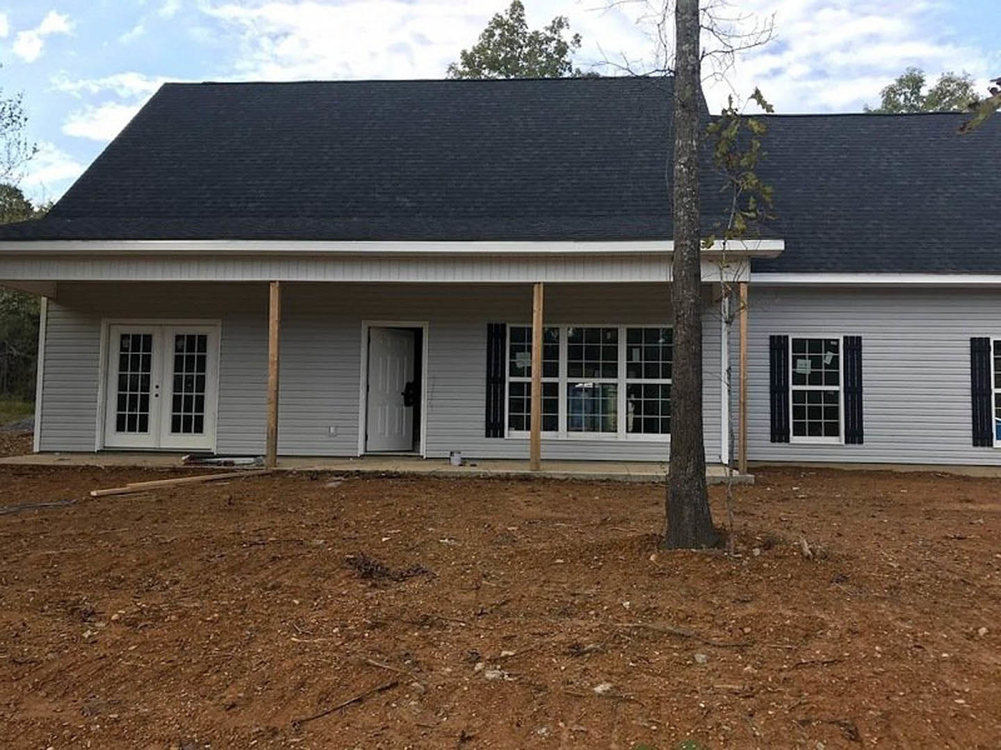 Front porch with white double doors featuring glass panes, black handles, and adjacent white garage; tree in background, dirt ground, gray roof, and cloudy sky.
