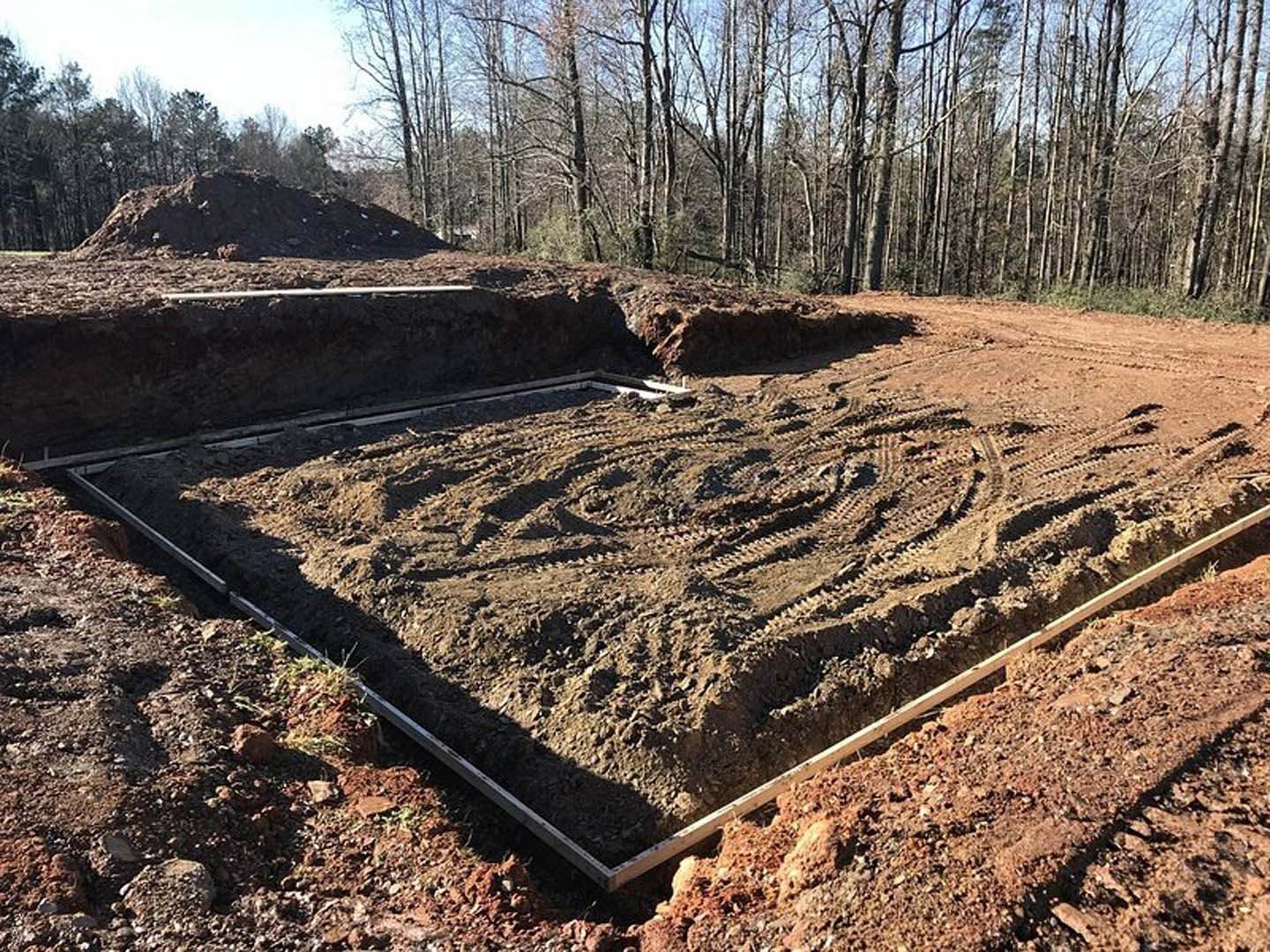 Dirt lot with excavated hole, tire tracks, wooden beam, metal pipe, and surrounding trees under open sky