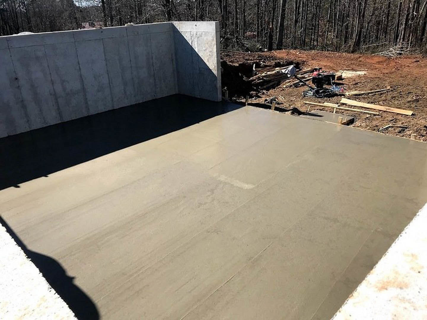 Freshly poured concrete slab with smooth finish, bordered by a concrete wall, surrounded by trees and wooded landscape, construction equipment visible in background, shadow of