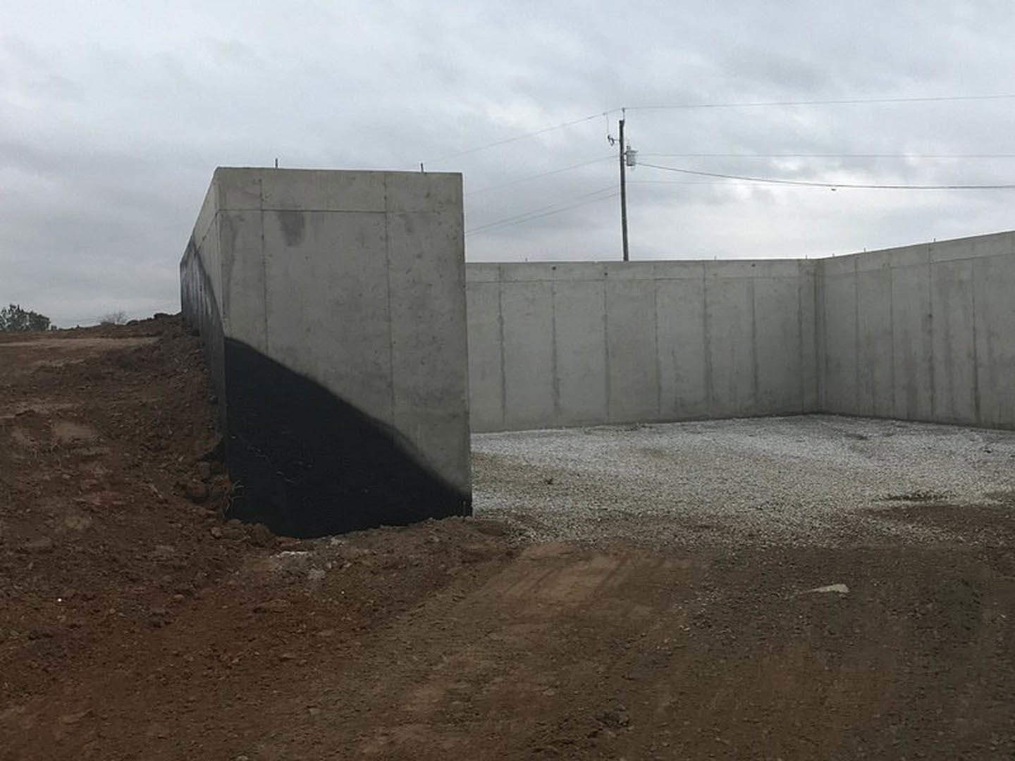 Black-painted concrete wall with vertical pillars, shadow cast across surface, distant trees visible beyond, grey carpet in foreground