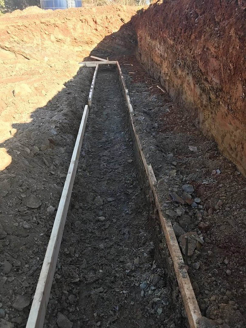 Wood beams and planks lining a dirt trench, white rectangular object partially buried, dirt walls and pile of rocks visible in foreground, outdoor construction site.