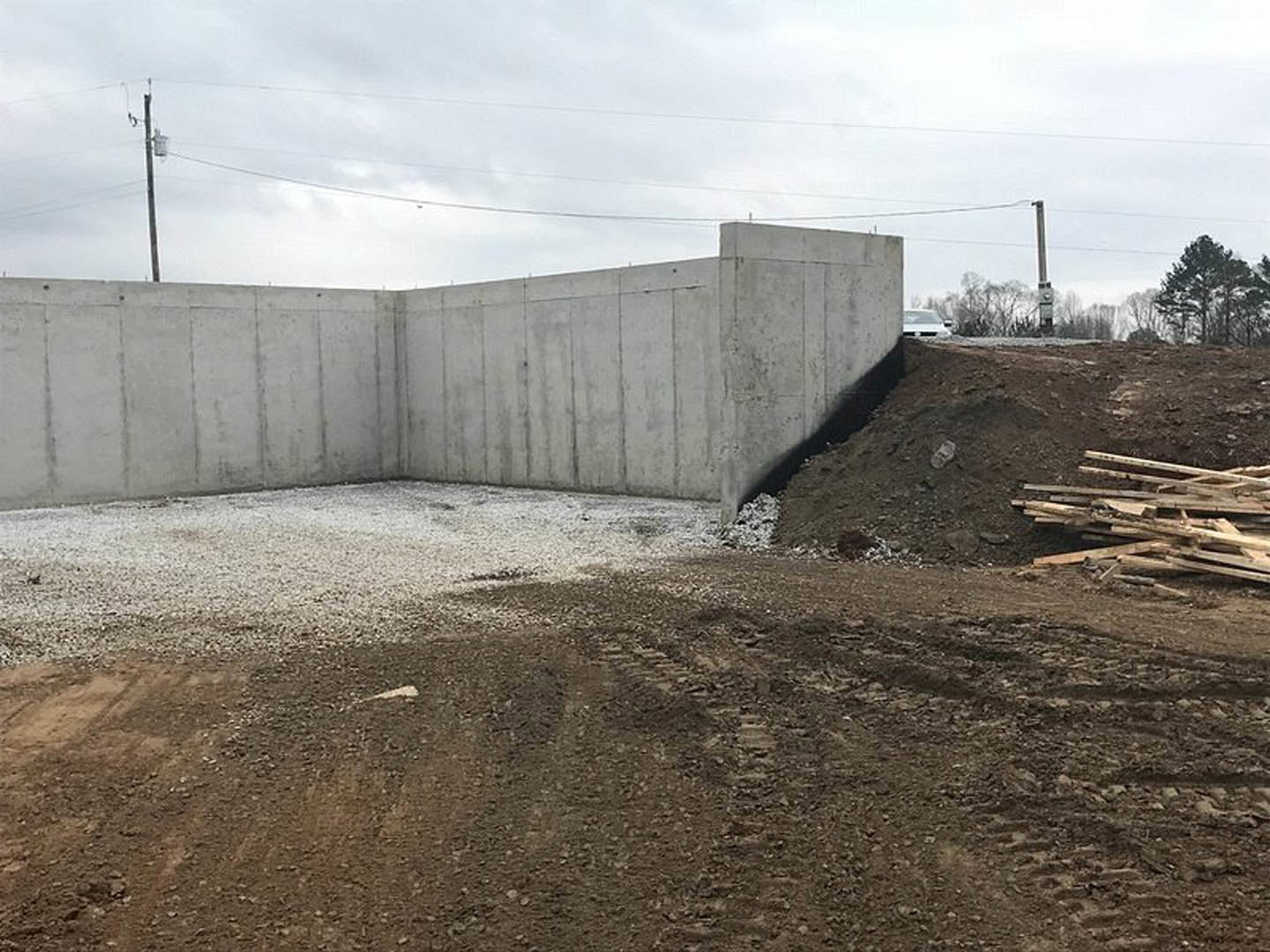 Concrete foundation wall beside exposed dirt and stacked wood pieces, power lines overhead, distant trees visible under cloudy sky