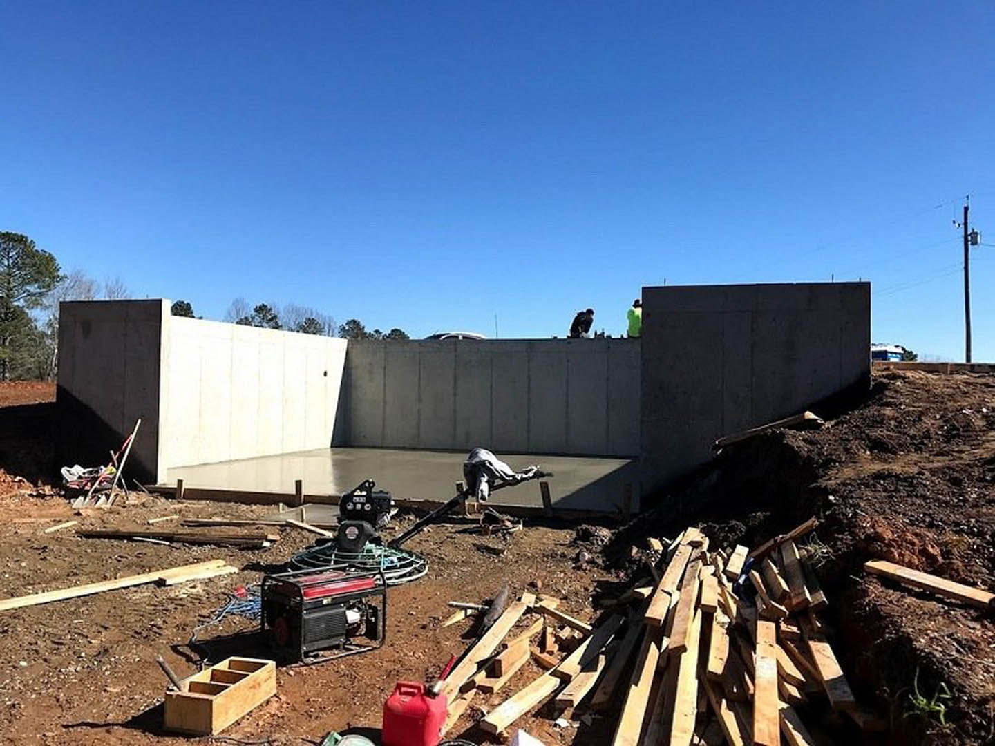 Workers building a concrete basement wall at a residential construction site, surrounded by dirt, scattered tools, a red gas can, and a wooden box with four compartments; trees and