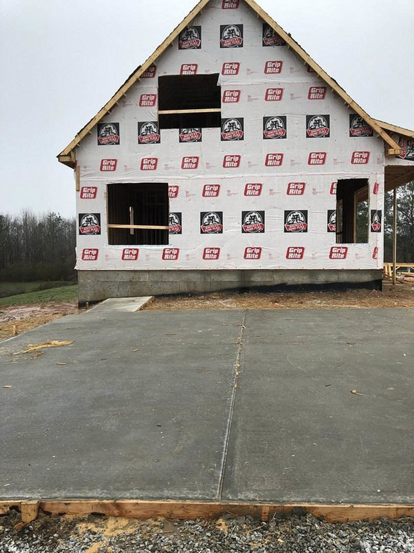 Partially built house with exposed wooden framing, completed roof, and freshly poured concrete driveway; surrounding area includes trees and construction materials.