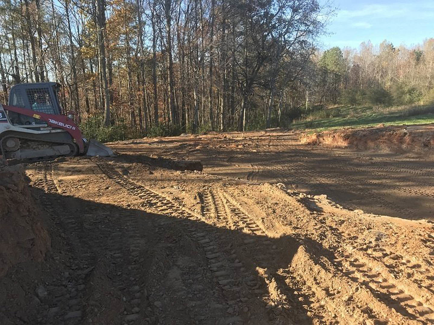 Tractor with large tires driving on a muddy dirt road beside a forested area, tire tracks visible in soil under a clear sky