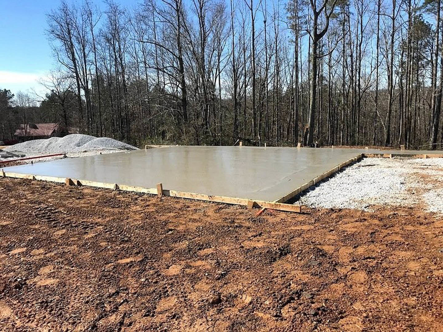 Freshly poured concrete slab foundation bordered by wooden forms, surrounded by dirt, gravel, and scattered trees under an open sky