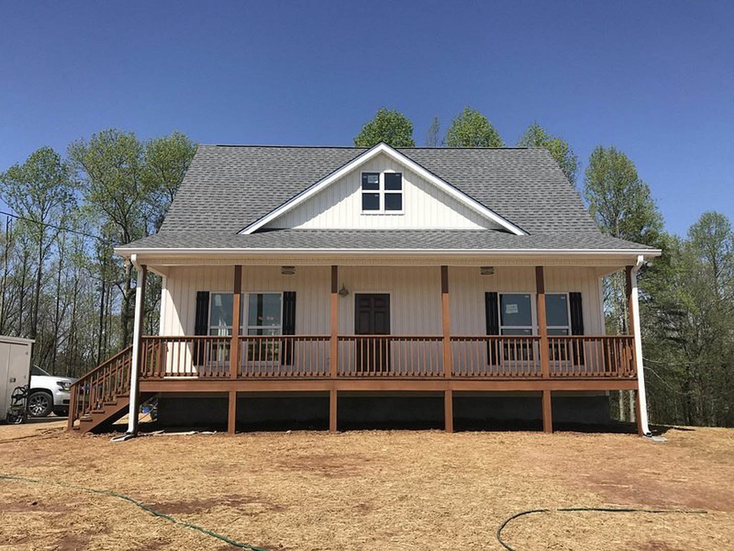 Two-story home with light siding, elevated wooden deck featuring metal railing, fenced backyard, large windows, white door with black handle, mature trees in background