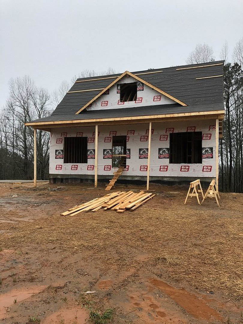 Framed house under construction with exposed wood planks, pile of lumber and sawhorses on dirt, surrounded by trees and sky