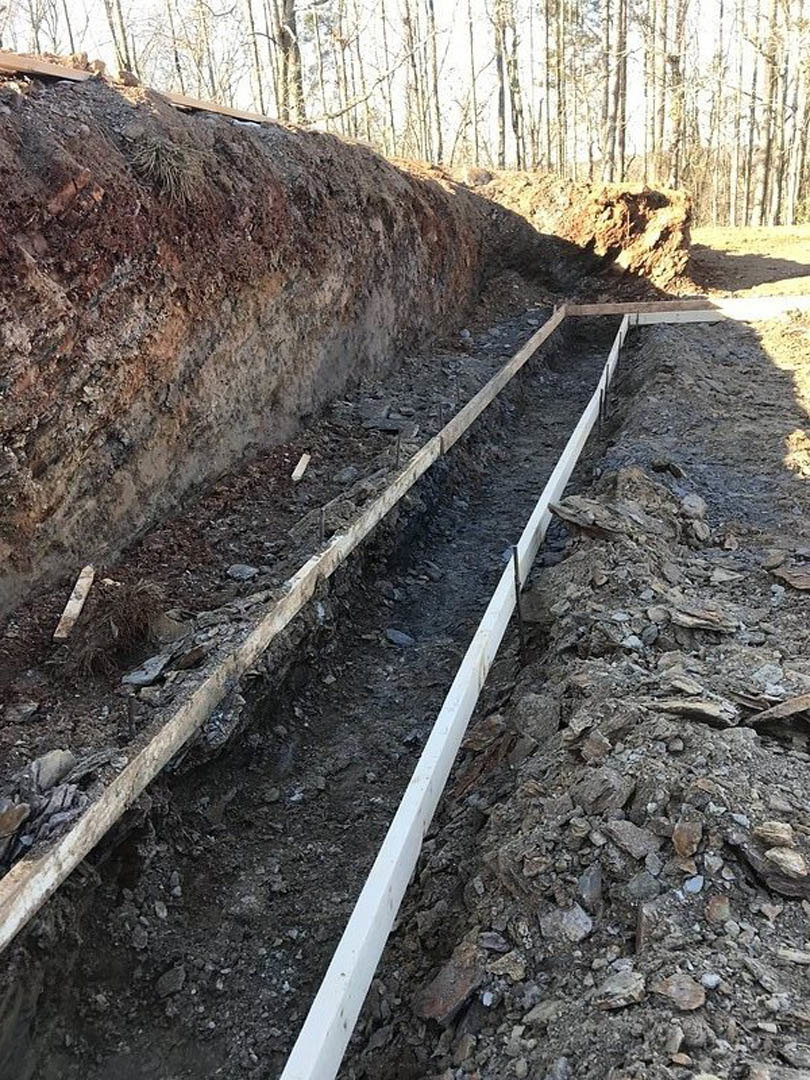 Deep trench dug into soil with wood planks lining the sides, white marking line on ground, pile of rocks and dirt nearby, trees and plants in background