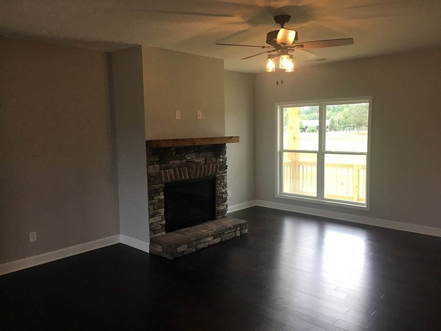 Living room with brick fireplace featuring a black insert and wood mantel, ceiling fan with lights, large window overlooking deck and trees, stone bench on black flooring