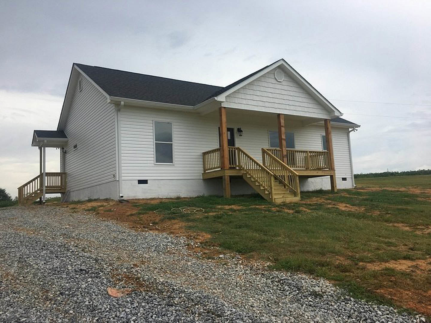 Two-story house with white-framed windows, wooden porch and stairs, gravel driveway bordered by grass, cloudy sky overhead