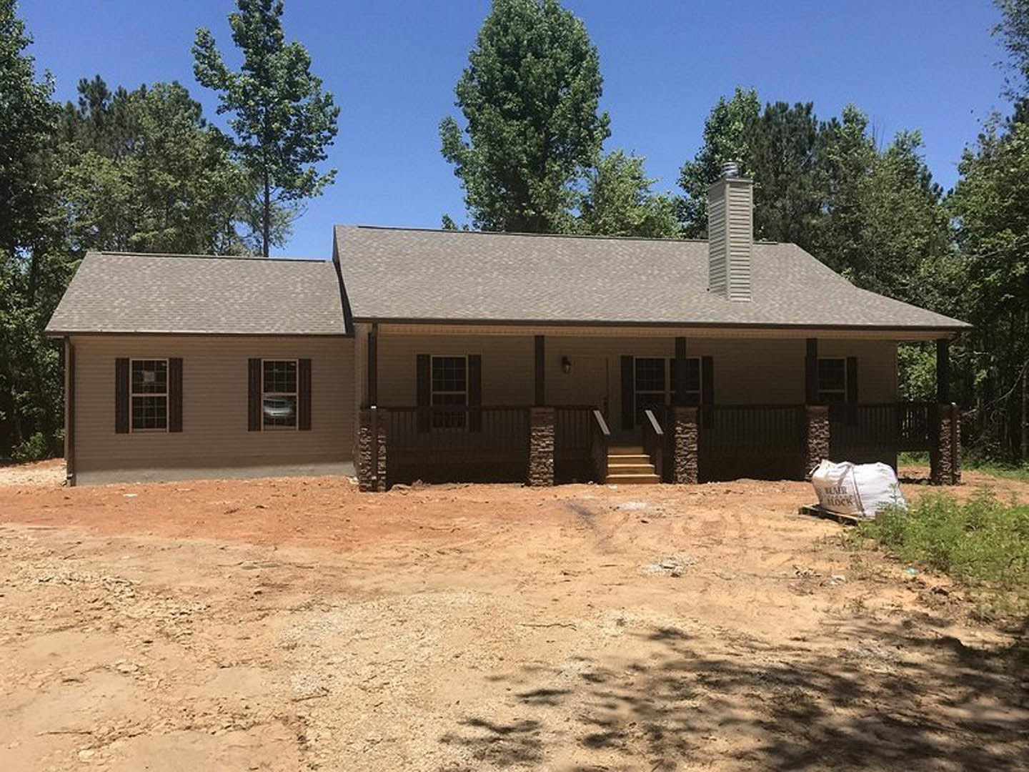 Single-story house with white siding, covered porch supported by square pillars, dirt yard in front, large white construction bag near entry, grid-style windows, parked car visible