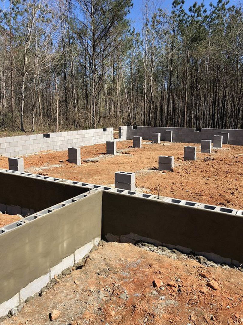 Concrete foundation slab with embedded blocks surrounded by soil, construction materials, and trees in the background