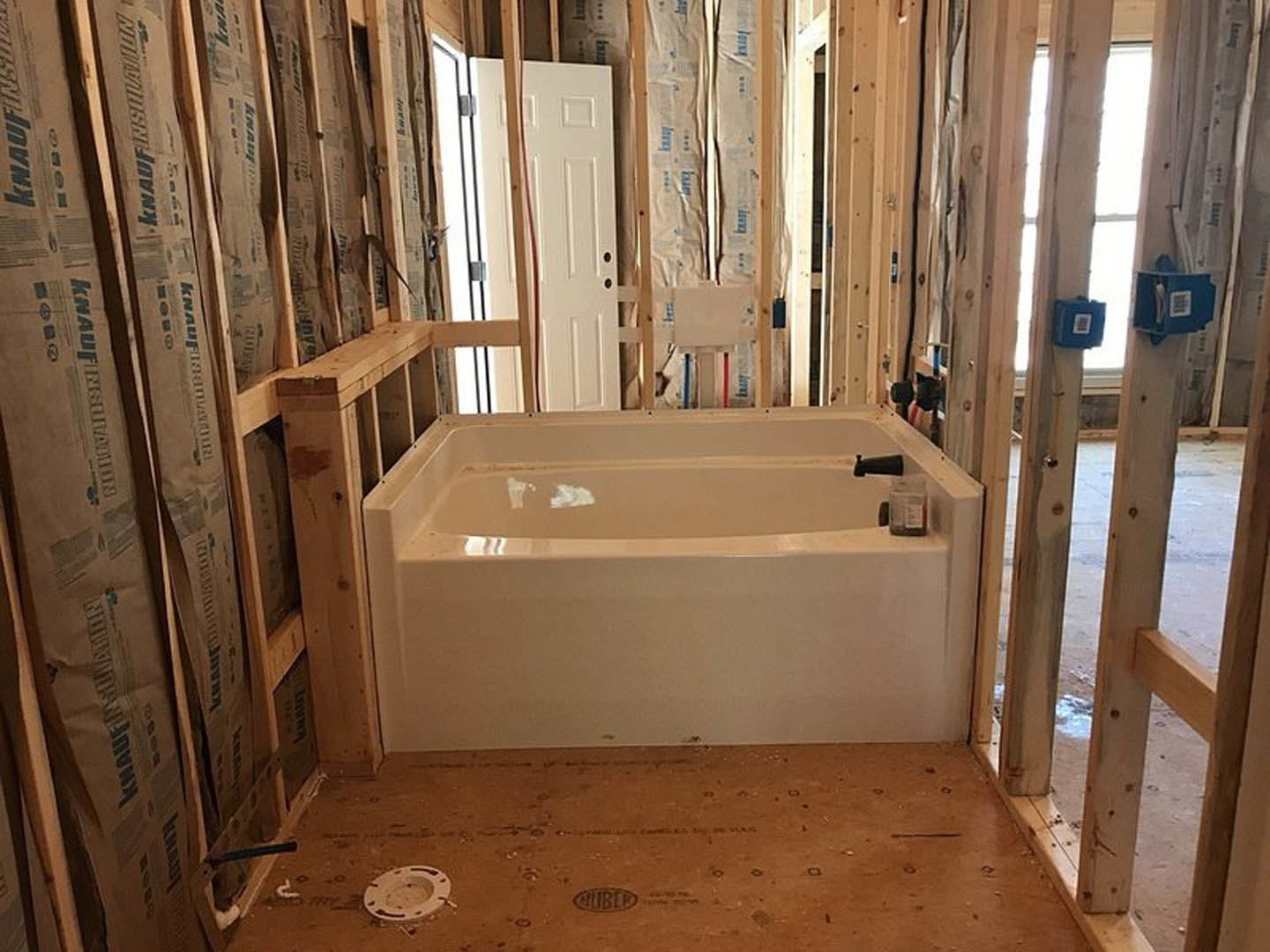 Freestanding white bathtub with chrome faucet against tiled wall, wood flooring, and large window in modern bathroom