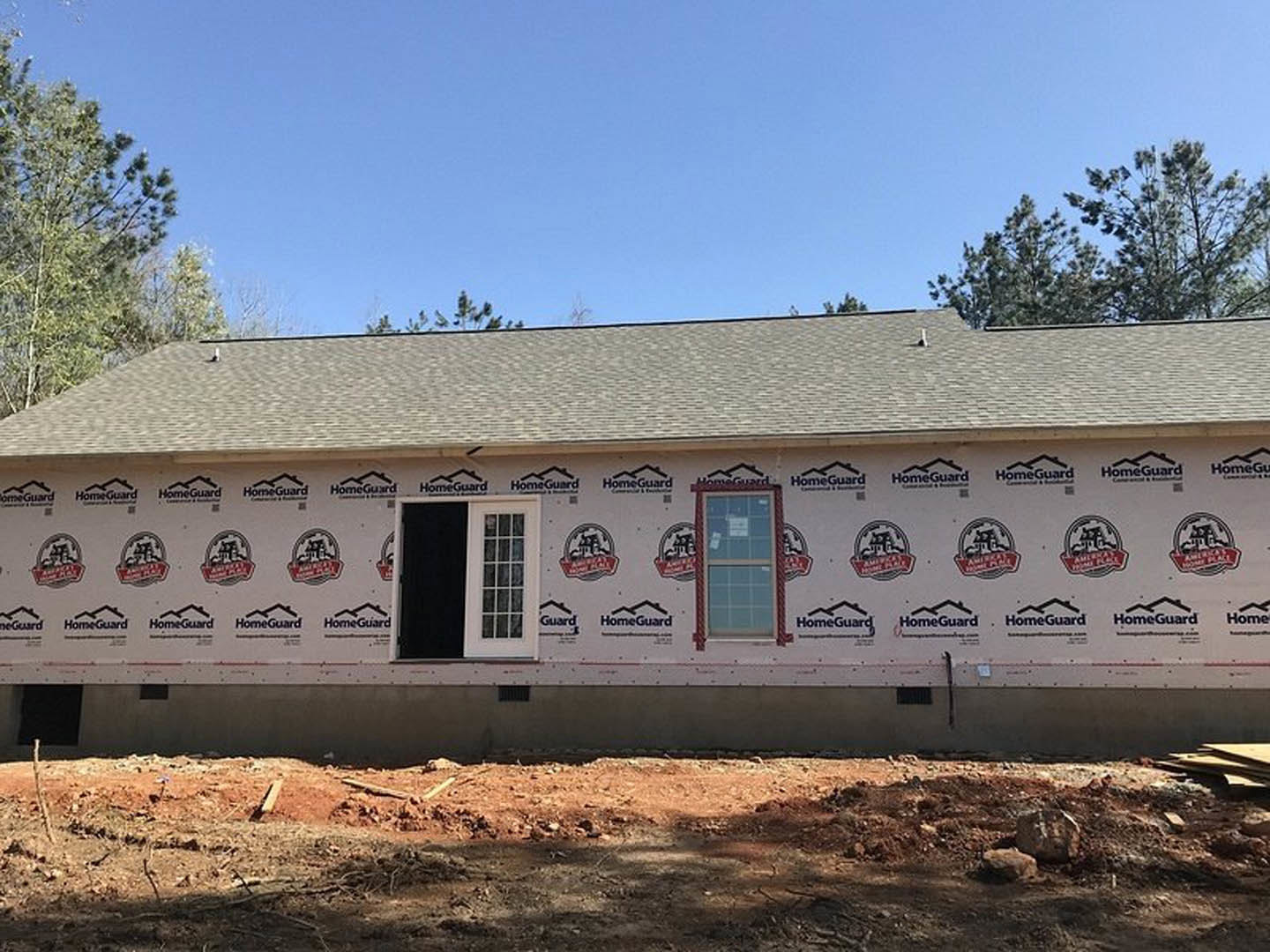 Exterior view of a custom home with one wall partially covered in construction materials, multi-pane window, exposed adhesive, dirt ground, and surrounding trees under a clear sky.