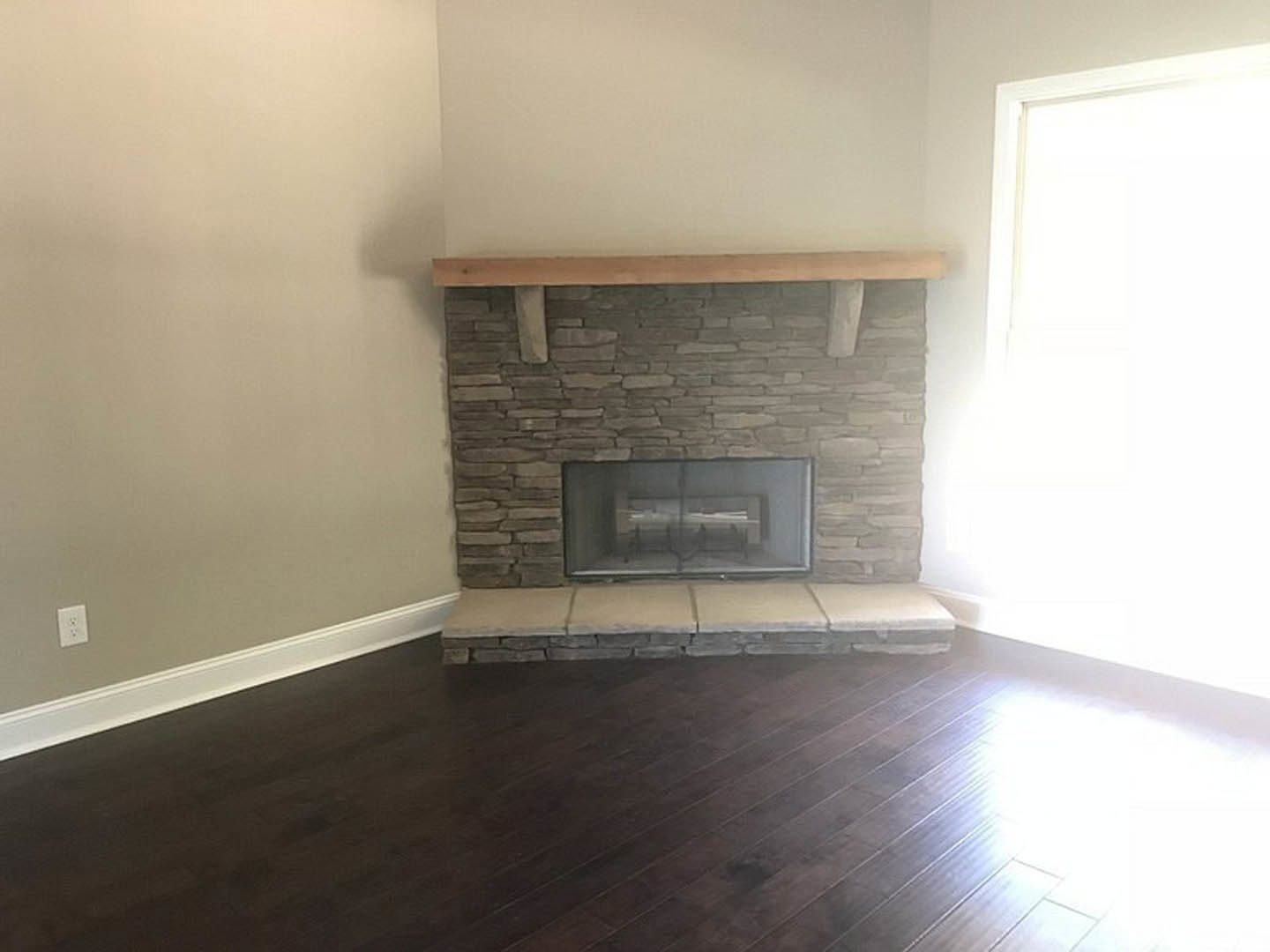 Stone fireplace with glass window, wood flooring, white plaster walls, and a white electrical outlet; person standing near window in a modern living room.