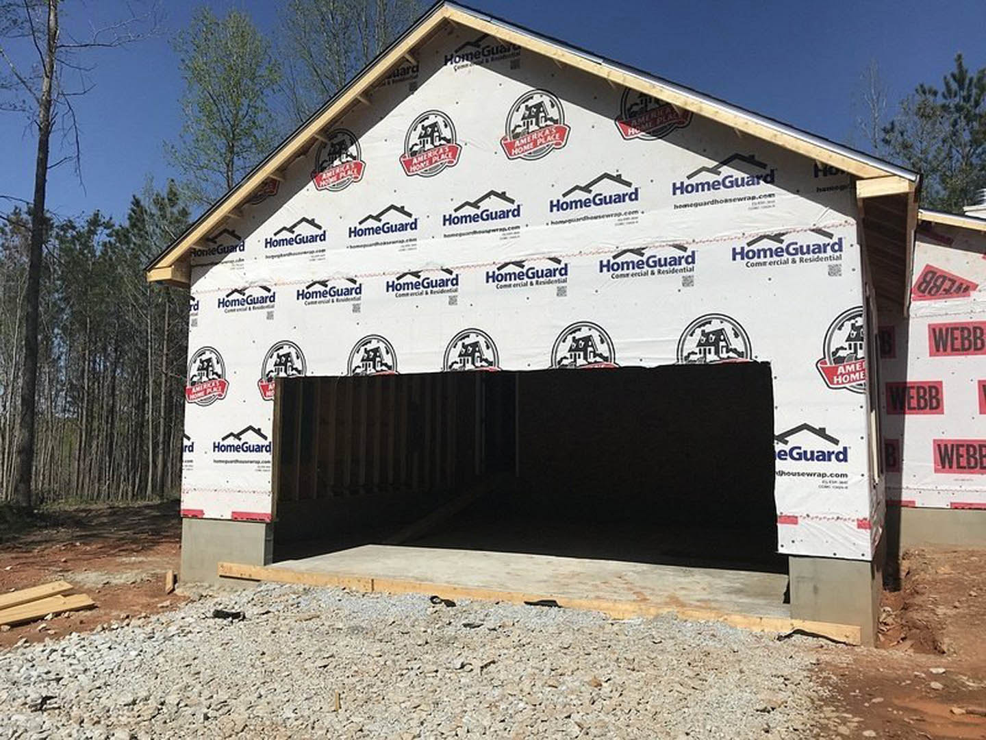 Partially constructed house with exposed framing, attached garage, gravel driveway, and surrounding trees under a clear sky