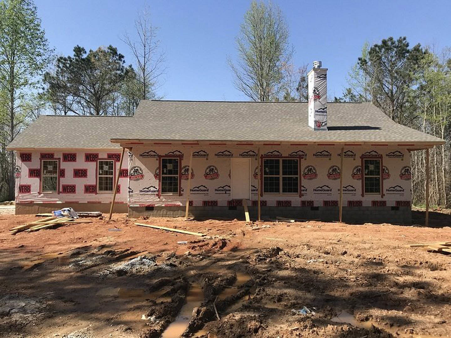 Two-story house under construction with gray roof, exposed framing, ladder leaning against exterior, muddy ground, scattered construction materials, trees in background, window