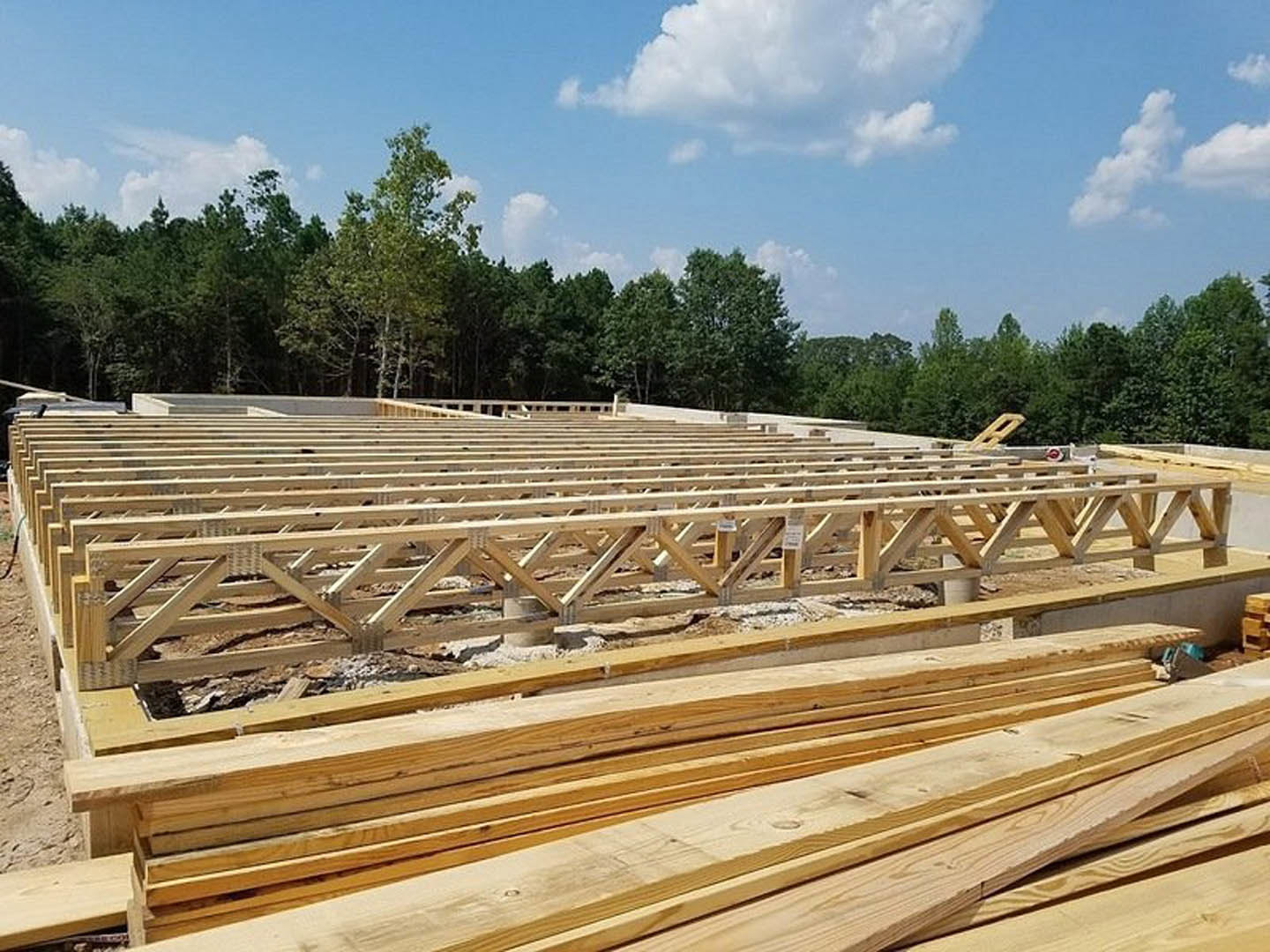 Exposed wooden beams and stacked lumber on a building exterior beneath a blue sky with scattered clouds and surrounding trees