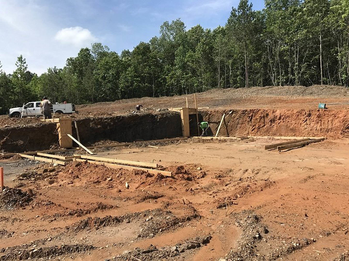 Framed foundation with exposed wooden beams on dirt ground, workers in safety gear near a white truck, trees bordering construction site under cloudy sky