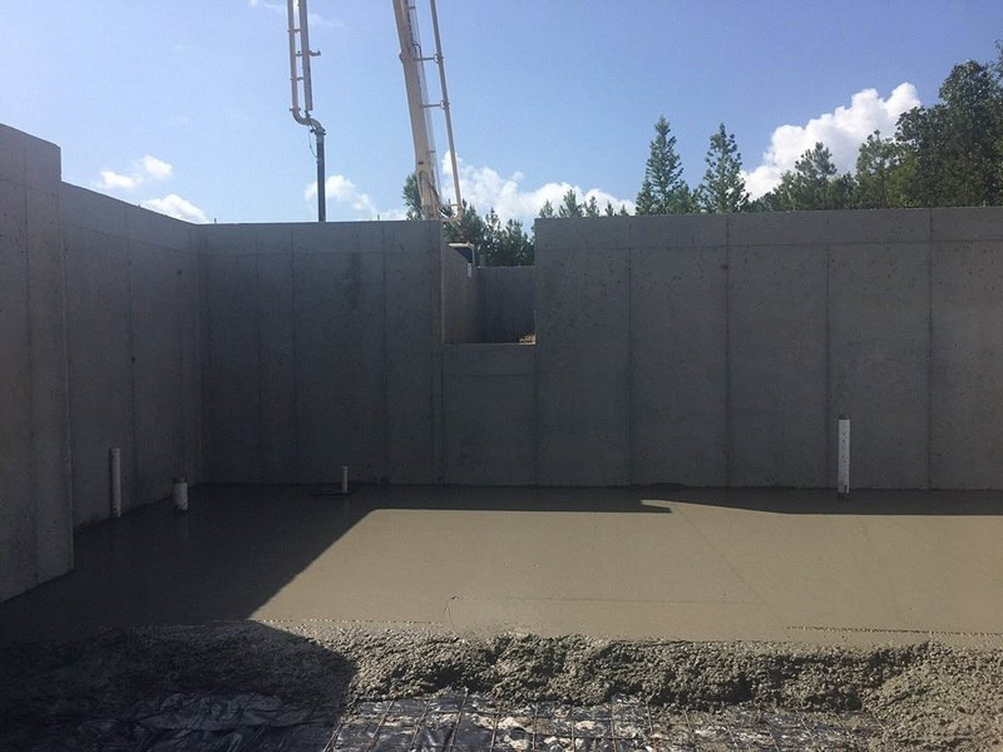 Concrete wall with trees and a person walking on the sidewalk, large metal crane and blue sky with clouds in the background, close-up of cement pile in foreground