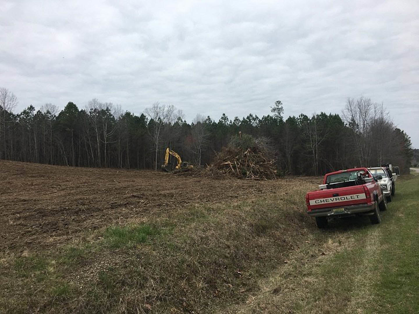 Red truck parked on grassy field near trees under cloudy sky, yellow excavator partially visible in wooded area