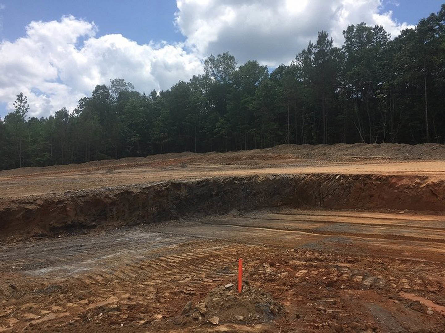 Dirt pit with a mound of soil and an orange pole at center, surrounded by trees under a cloudy sky