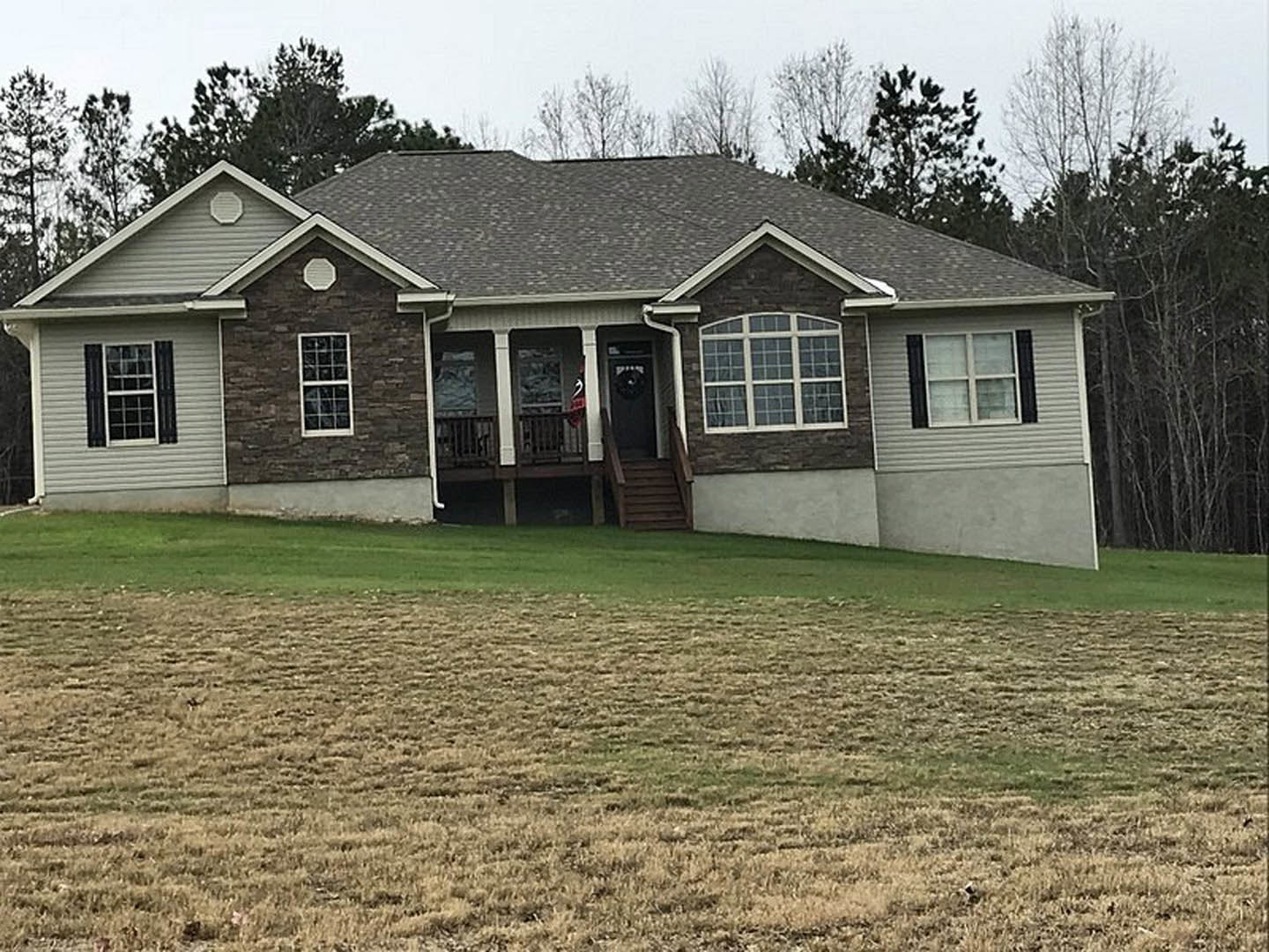 White farmhouse-style home with black shuttered windows, covered front porch, wooden stairs, and manicured green lawn bordered by mature trees