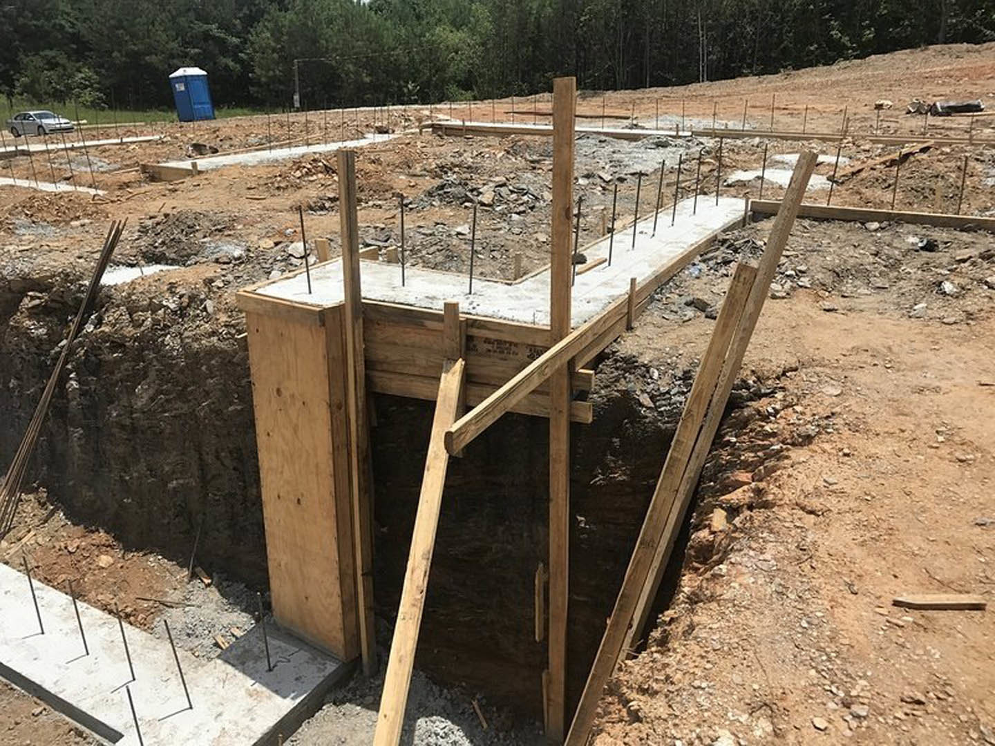 Concrete foundation with exposed metal rods set in soil, surrounded by construction materials, portable toilet, and trees in the background