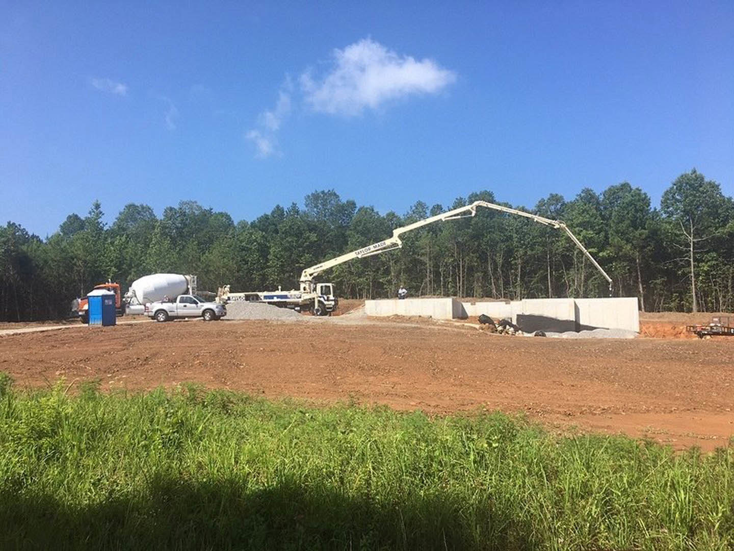 White truck with crane lifting concrete slab above partially built wall, open truck door visible, grassy dirt field in foreground, scattered clouds in blue sky.