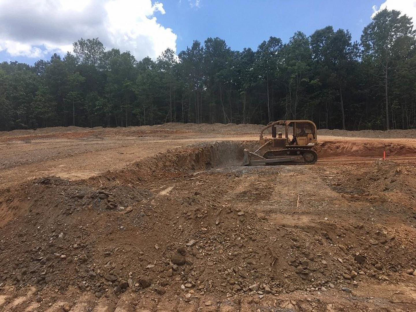 Bulldozer excavating soil in a dirt field bordered by trees under a cloudy sky