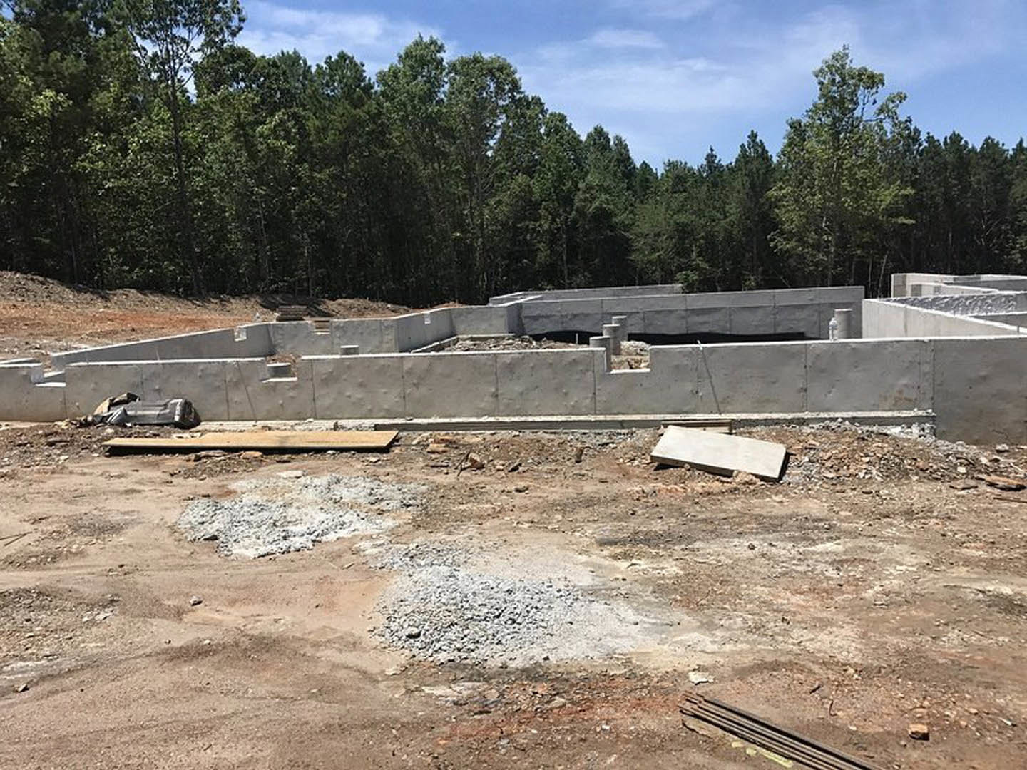 Concrete foundation with exposed rebar and gravel piles, unfinished walls, white board on ground, surrounded by trees under cloudy sky