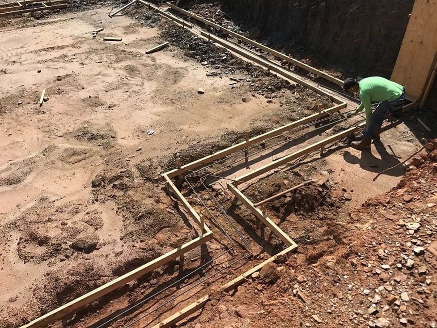 Wooden framing and metal rebar set in soil at a residential construction site, with a man in a green shirt working near the foundation.