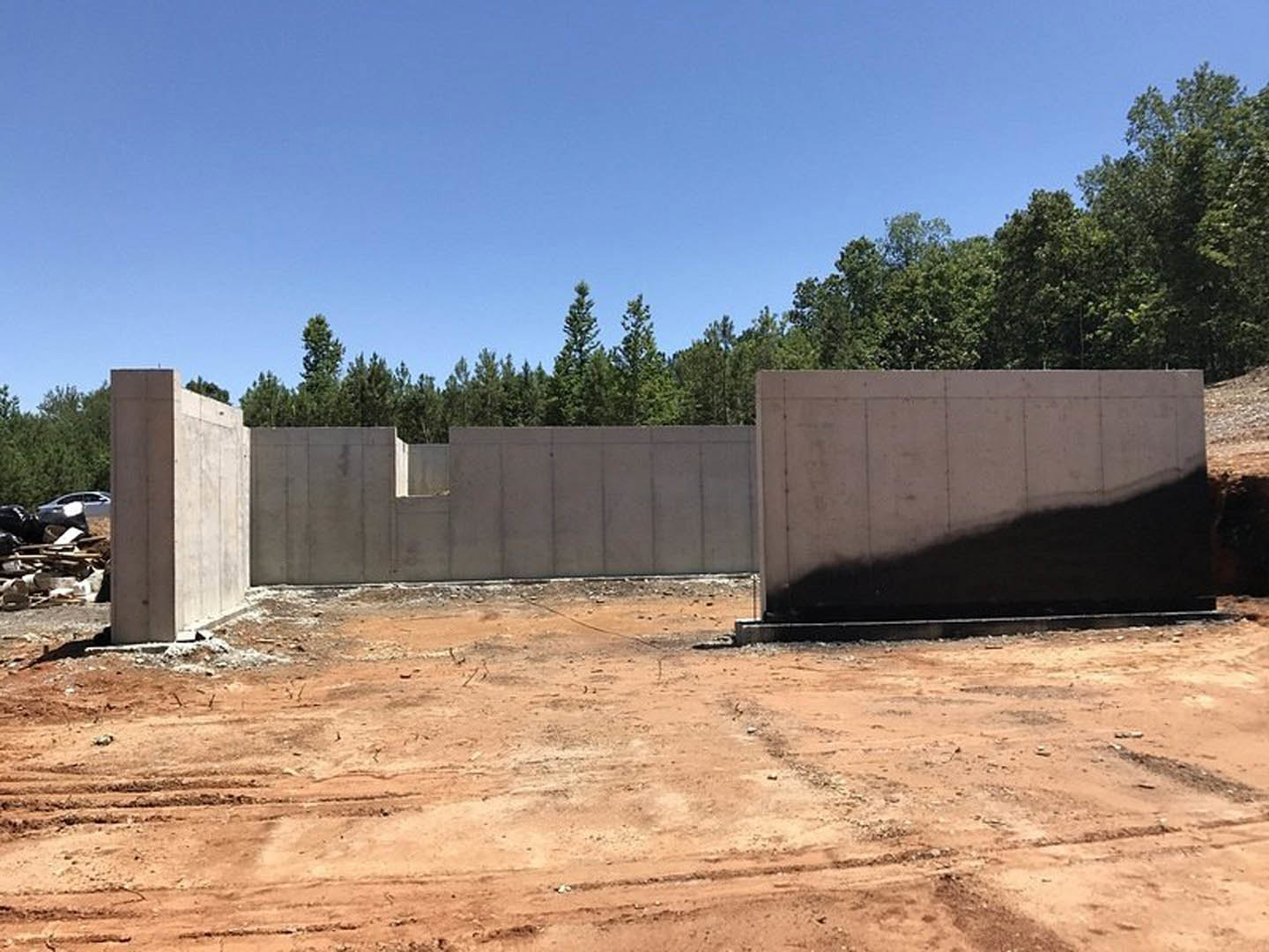 Grey concrete wall with a shadow cast by a person, dirt ground with a metal pole, trees and blue sky in the background