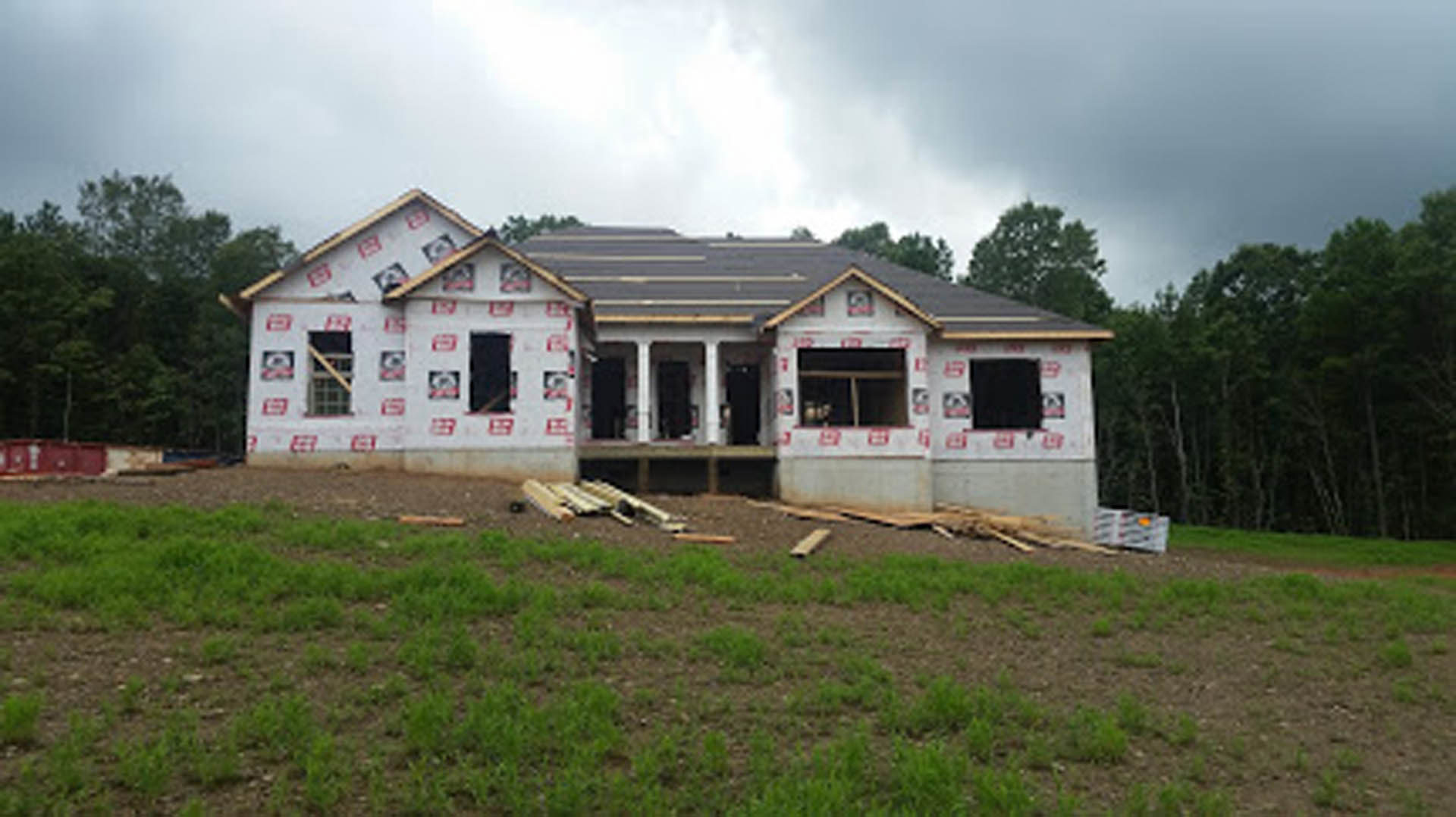 Partially built house with exposed wooden framing, concrete pillars, and unfinished porch surrounded by grass and scattered lumber, mature trees in background under cloudy sky
