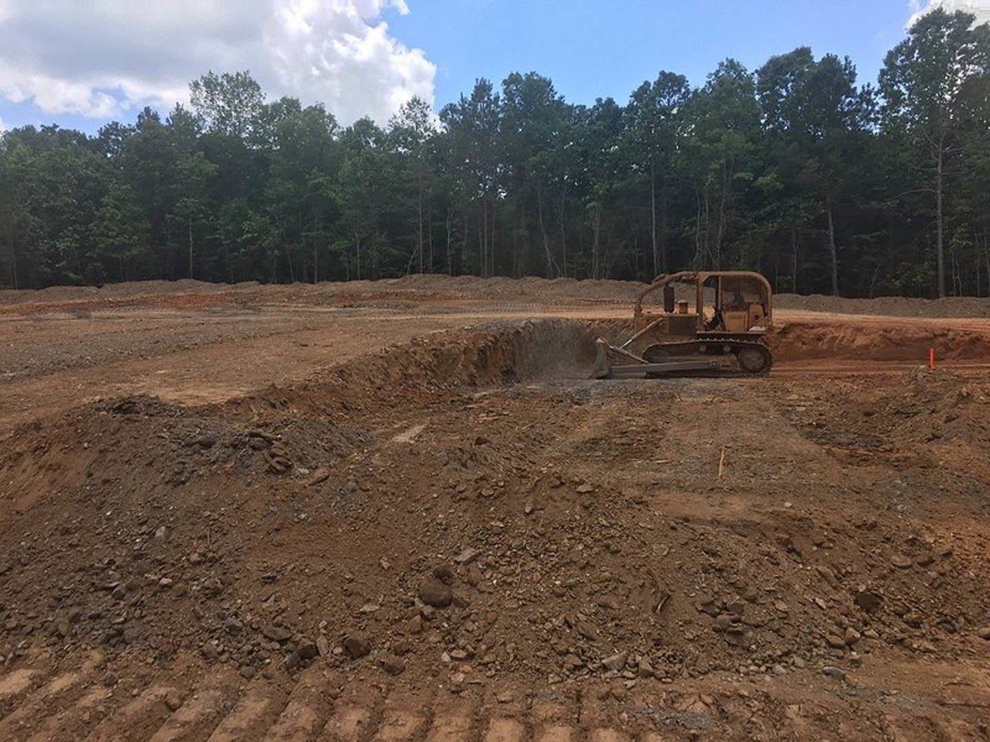 Yellow bulldozer grading soil on a dirt field with scattered trees and cloudy sky in the background