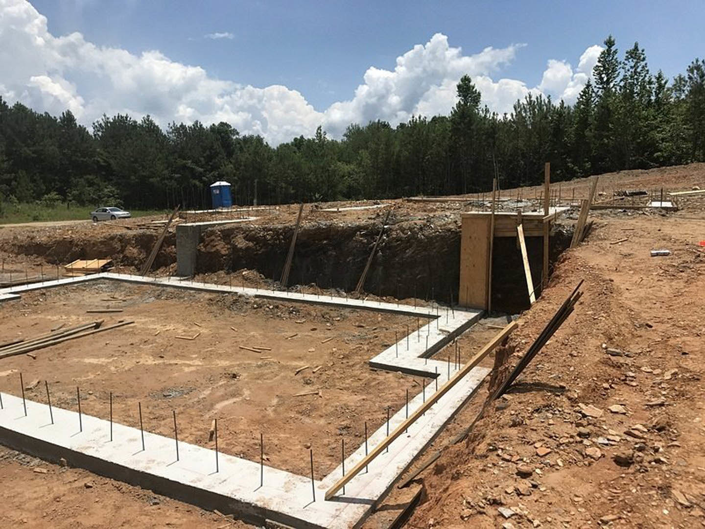 Concrete foundation with exposed metal rods surrounded by soil, wooden beam structure partially built, blue portable toilet near trees, sky with scattered clouds overhead
