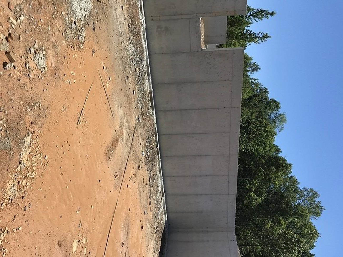 Concrete exterior wall under construction, dirt path and rocky ground in foreground, mature trees with green leaves in background