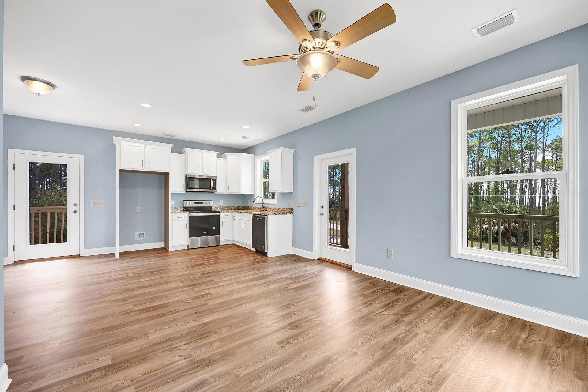 Open living area with hardwood floors, ceiling fan with light fixture, large window overlooking deck with bird on railing, kitchen stove visible, door opens to outdoor view of