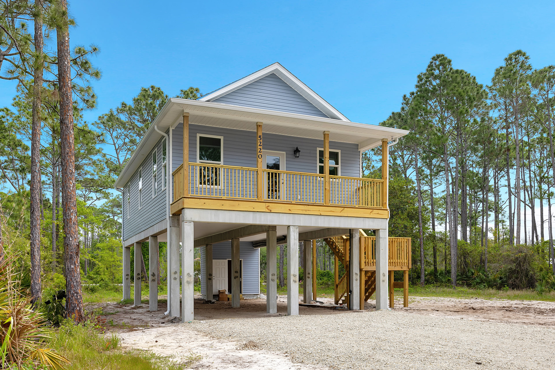 Elevated house on wooden stilts with a covered porch, exterior staircase, and walkway, surrounded by trees and plants, featuring large windows and a garage underneath