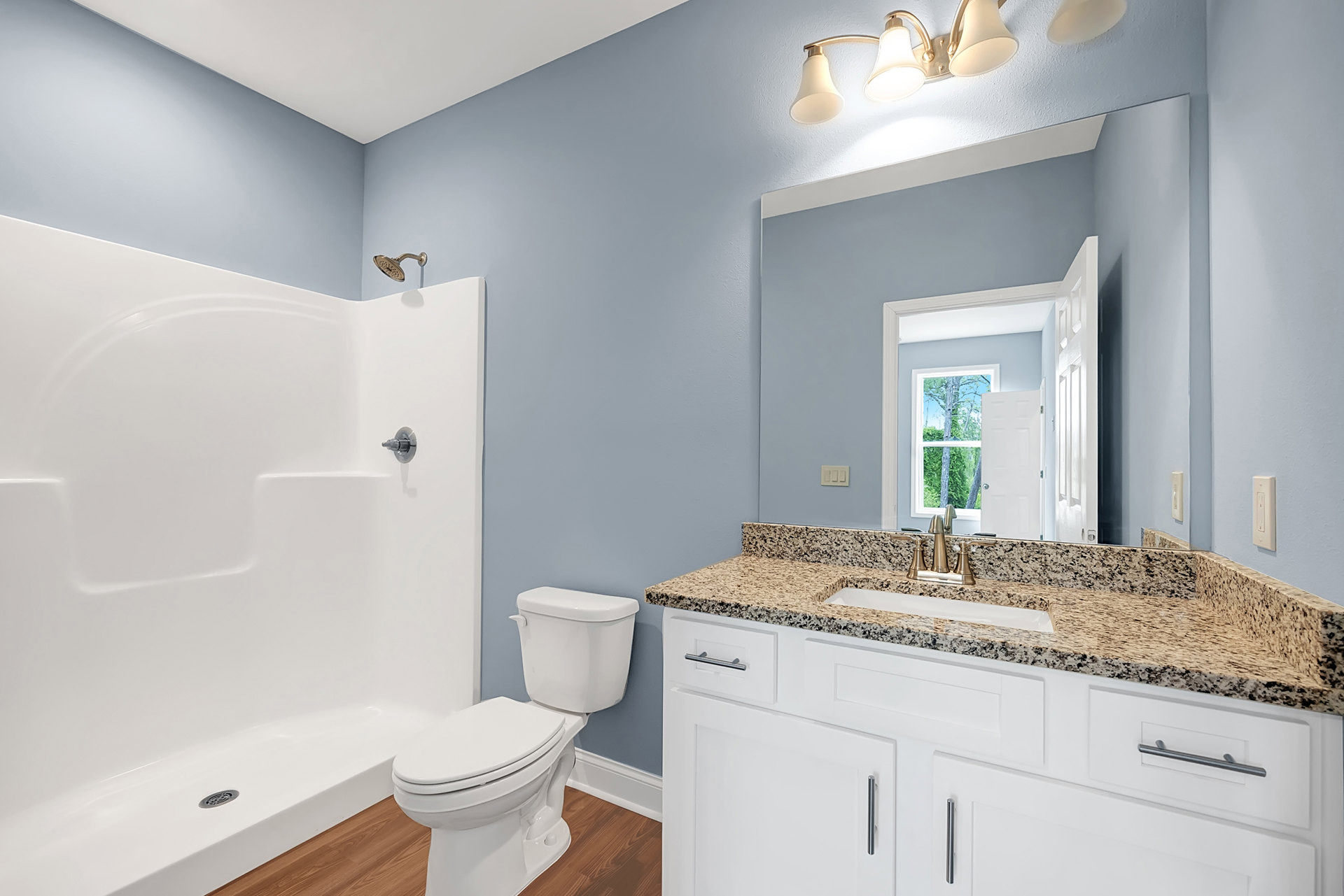 White toilet and glass-enclosed shower on wood floor, white tile walls, double light fixture above sink with stone countertop and cabinetry